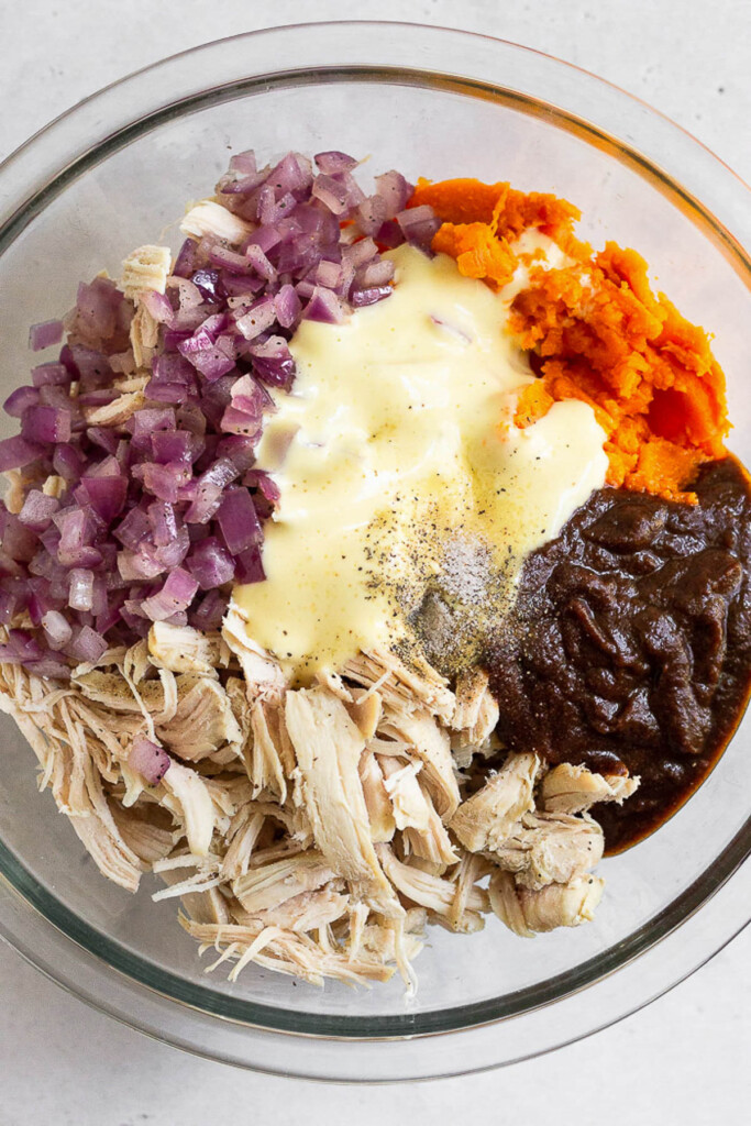 Glass bowl filled with mashed sweet potato, barbecue sauce, shredded chicken, sautéed red onion, and mayo before it is mixed together.