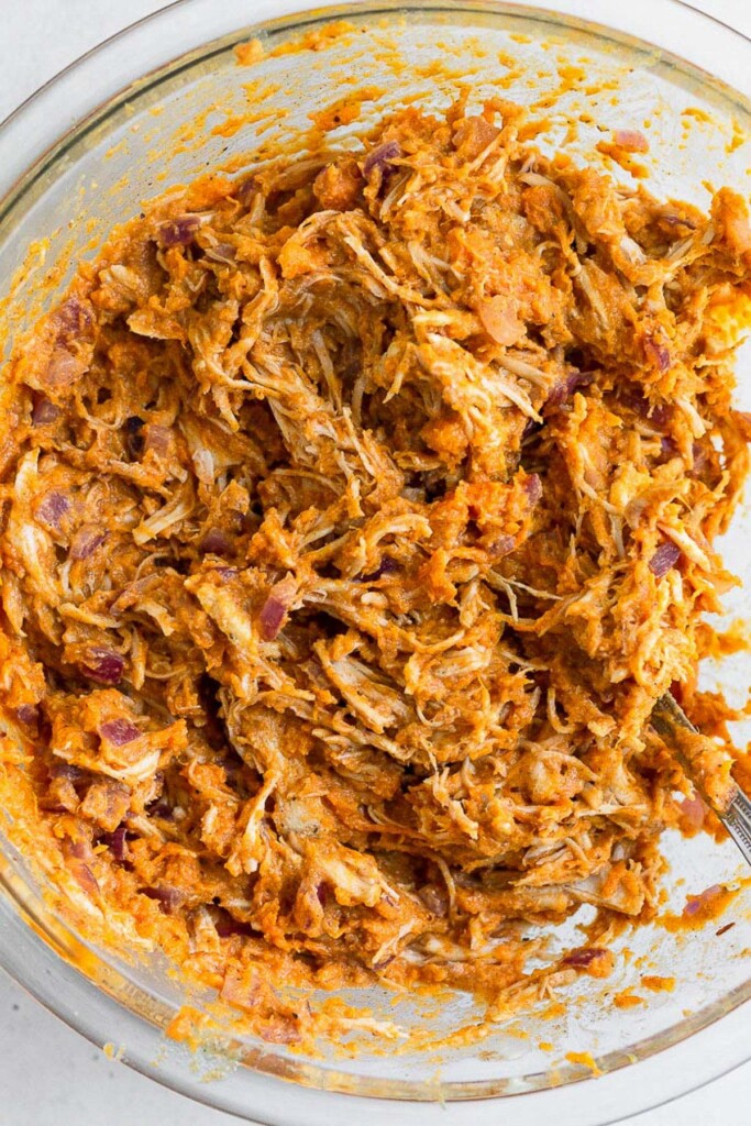Large glass bowl filled with shredded chicken mixed with cooked mashed sweet potato, sautéed onions, and spices.