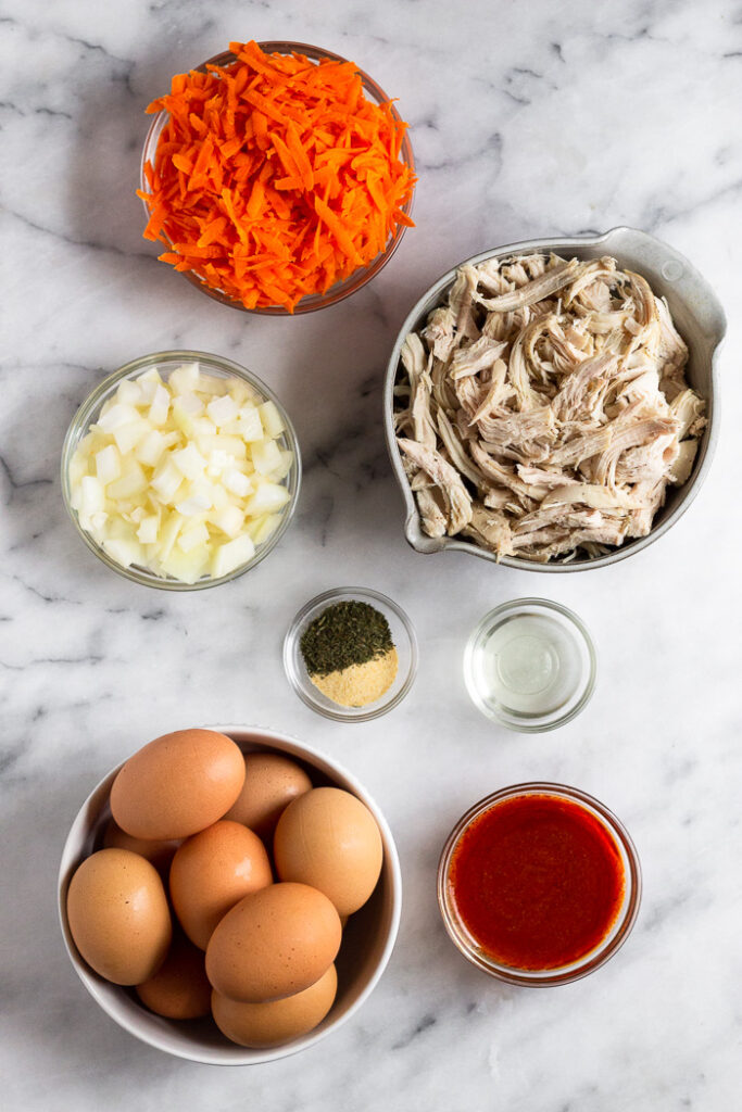 Overhead shot of a bowl filled with shredded carrots, bowl filled with shredded chicken, bowl of oil, bowl of buffalo sauce, bowl of eggs, bowl of spices, and a bowl of diced onion.