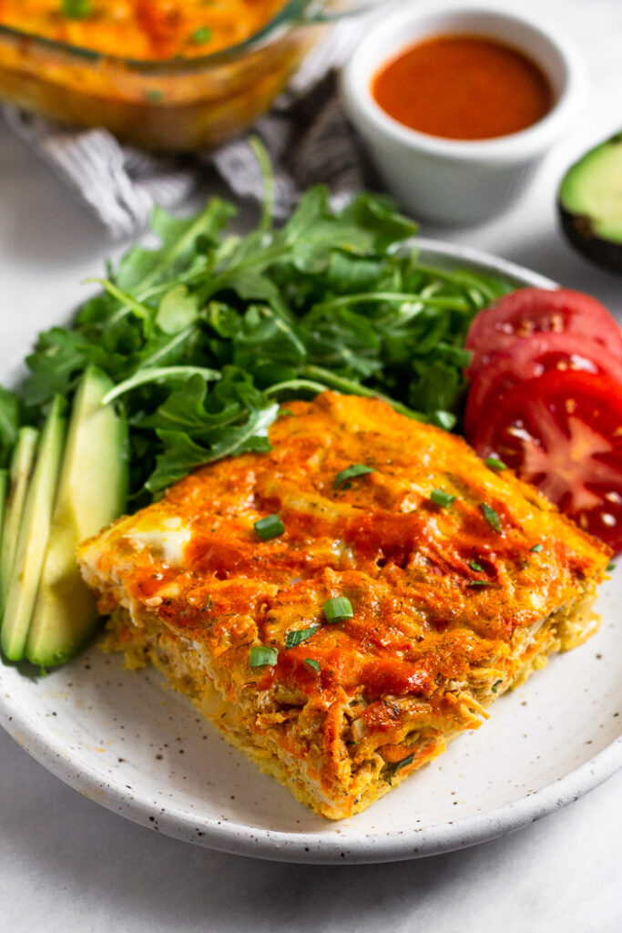A piece of paleo breakfast casserole on a plate with sliced avocado, green, and sliced tomato. Behind it is a large dish of the rest of the casserole and a small bowl of buffalo sauce.