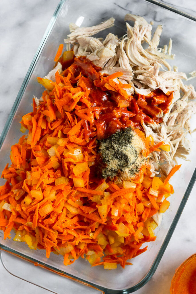 Glass casserole dish filled with sautéed shredded carrots, diced onions, spices, shredded chicken, and buffalo sauce. Everything isn't mixed together.