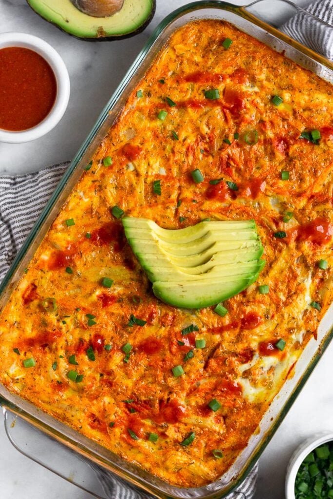 Overhead shot of a low carb buffalo chicken breakfast casserole in a large baking dish. It is topped with sliced avocado, hot sauce, and green onions. Next to is a half an avocado, small dish of buffalo sauce, and a small bowl of green onions.