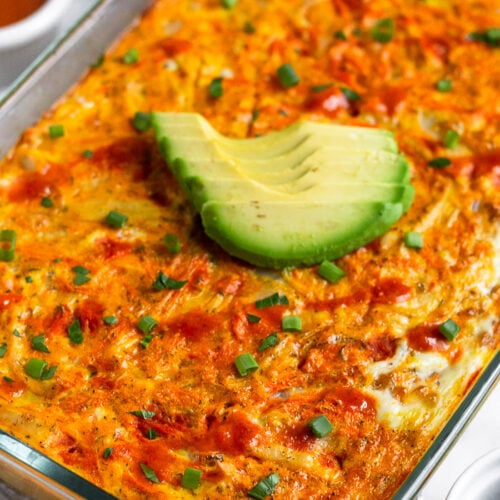 Chicken breakfast casserole in a casserole dish topped with avocado, hot sauce, and green onions. Next to it a ramekin of chopped green onions, ramekin of buffalo sauce, and half an avocado.