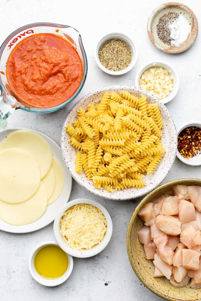 Overhead shot of a measuring cup of pasta sauce, a bowl of spices, a bowl of salt and pepper, a bowl of minced garlic, a bowl of uncooked fusilli pasta, a bowl of red pepper flakes, a bowl of diced raw chicken, a bowl of olive oil, a bowl of parmesan cheese, and a plate of sliced cheese.