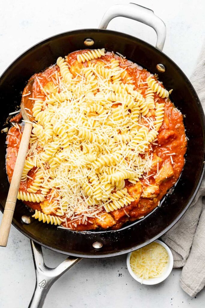 Skillet filled with cooked diced chicken mixed with marinara sauce. On top of that is a pile of cooked pasta and shredded parmesan cheese.