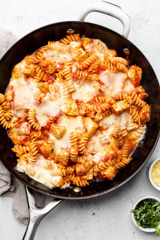 Skillet chicken parmesan with pasta in a large skillet. Next to it is a small bowl of chopped basil and a small bowl of parmesan cheese.