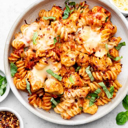 Chicken parmesan pasta in a bowl topped with fresh basil. Next to the bowl is more fresh basil, a bowl of red pepper flakes, and a bowl of parmesan cheese.