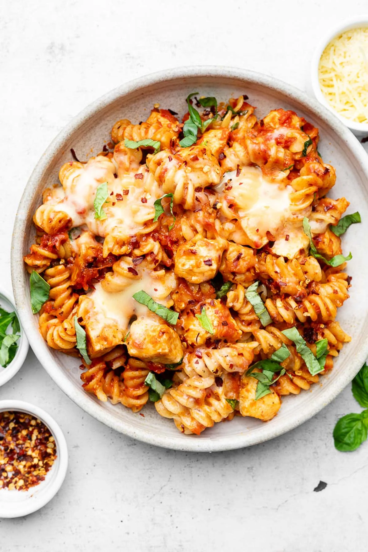 Chicken parmesan pasta in a bowl topped with fresh basil. Next to the bowl is more fresh basil, a bowl of red pepper flakes, and a bowl of parmesan cheese.