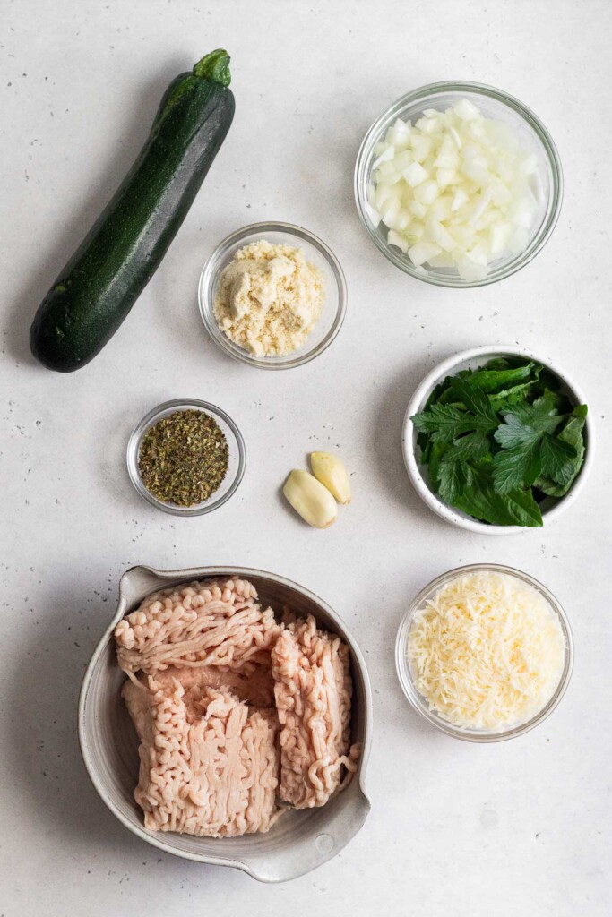 White counter with a bowl of diced onions, a bowl of fresh herbs, a bowl of grated parmesan cheese, a bowl of raw ground chicken, 2 peeled garlic cloves, a bowl of dried herbs, a bowl of flour, and a whole zucchini.