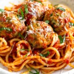 A bowl of zucchini meatballs on top of pasta mixed with marinara sauce. They are garnished with fresh basil and grated cheese.
