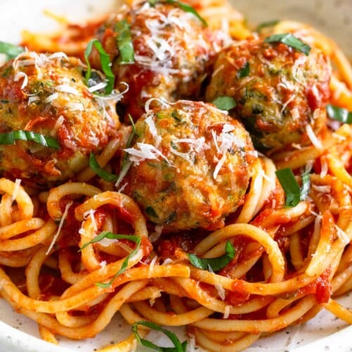 A bowl of zucchini meatballs on top of pasta mixed with marinara sauce. They are garnished with fresh basil and grated cheese.