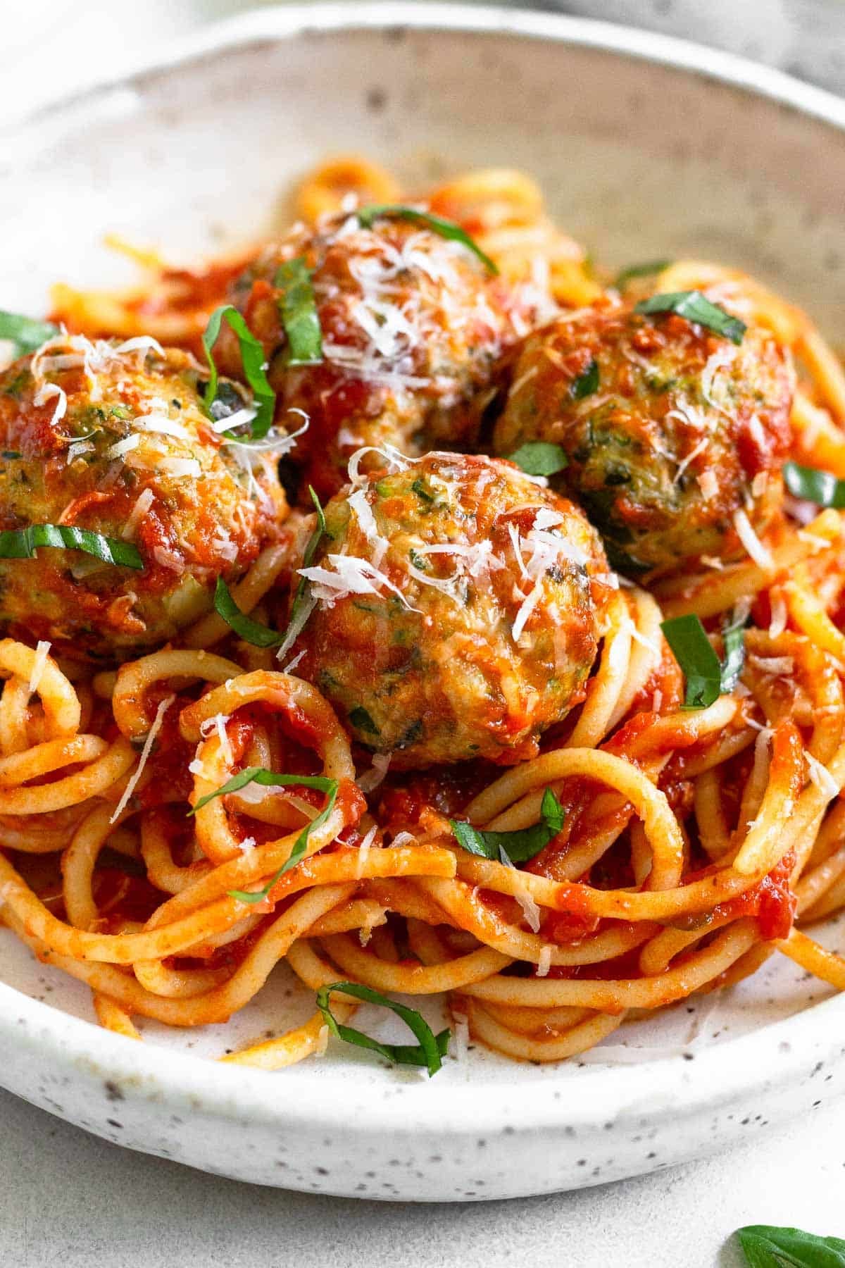 A bowl of zucchini meatballs on top of pasta mixed with marinara sauce. They are garnished with fresh basil and grated cheese.