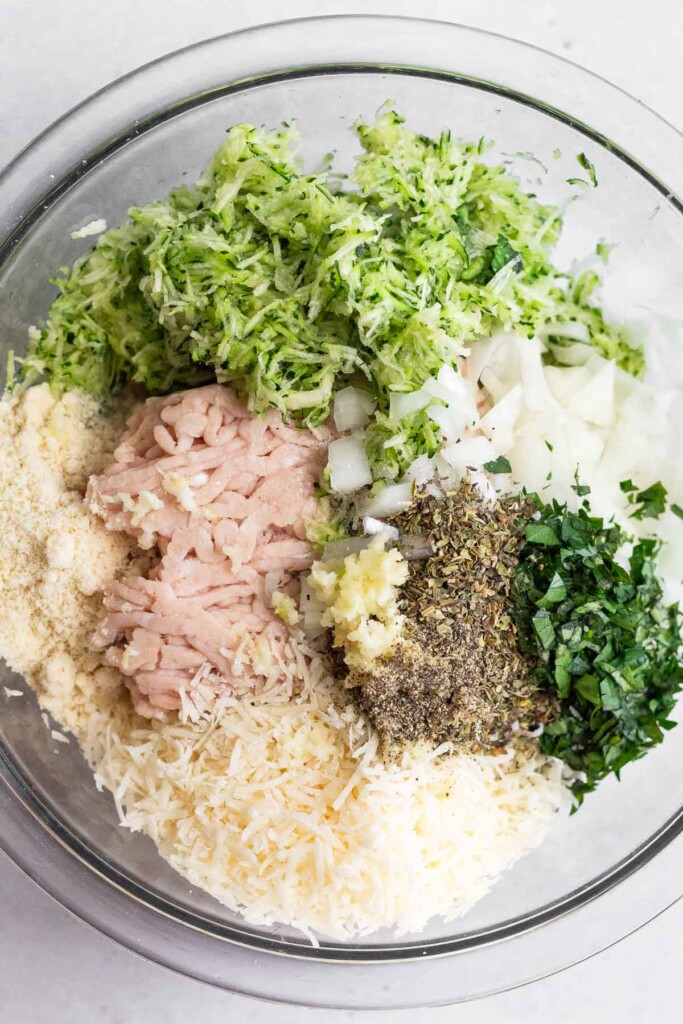 Large glass bowl filled with raw ground chicken, almond flour, shredded zucchini, diced onion, fresh herbs, dried herbs, parmesan cheese, and minced garlic.