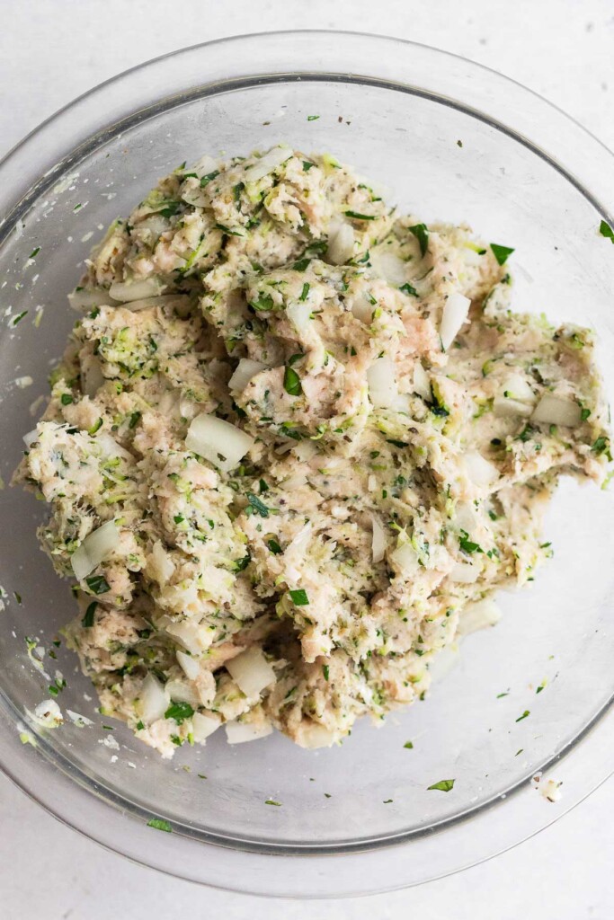 A bowl of raw ground chicken mixed with shredded zucchini, diced onions, herbs, and spices.
