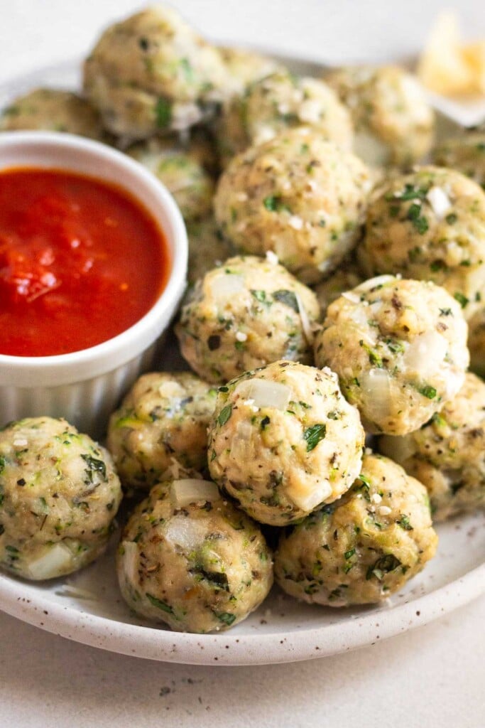 A plate of chicken zucchini meatballs stacked on top of each other with a ramekin of marinara sauce next to them.