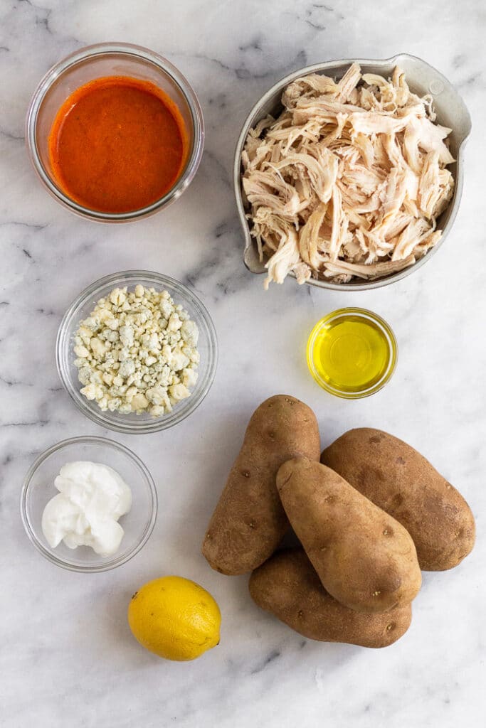 Marble countertop with a jar of buffalo sauce, bowl of shredded chicken, bowl of oil, 4 russet potatoes, lemon, small bowl of yogurt, and a bowl of blue cheese crumbles.