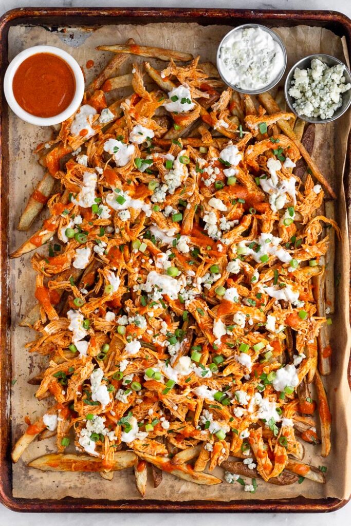 Large baking sheet with buffalo fries topped with buffalo chicken, blue cheese dressing, blue cheese crumbles, green onion, and parsley. There is also three small ramekins on the baking sheet, one has buffalo sauce, one has blue cheese dressing, and one with blue cheese crumbles in it.