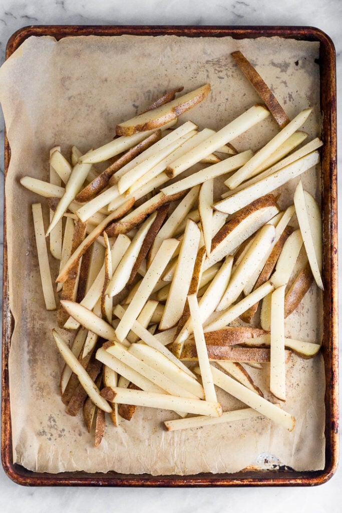 Overhead shot of raw French fries piled on a baking sheet lined with parchment paper.