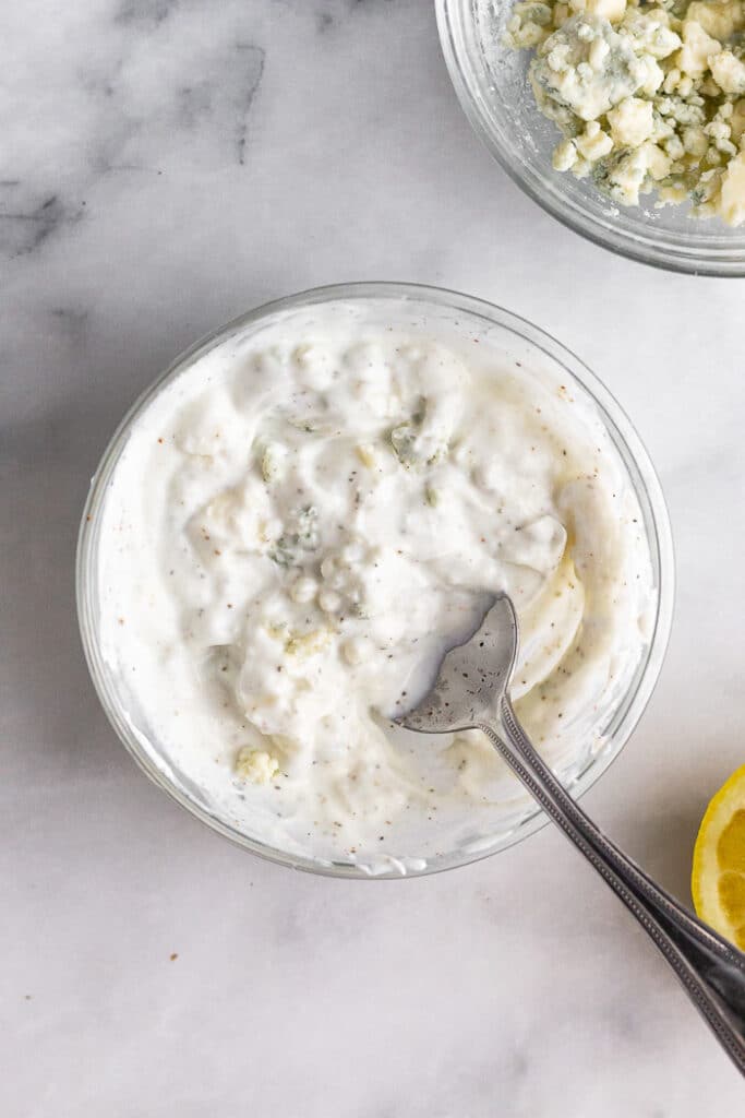 Small bowl of homemade blue cheese dressing with a spoon in it. Next to it is half a lemon and a bowl of blue cheese crumbles.