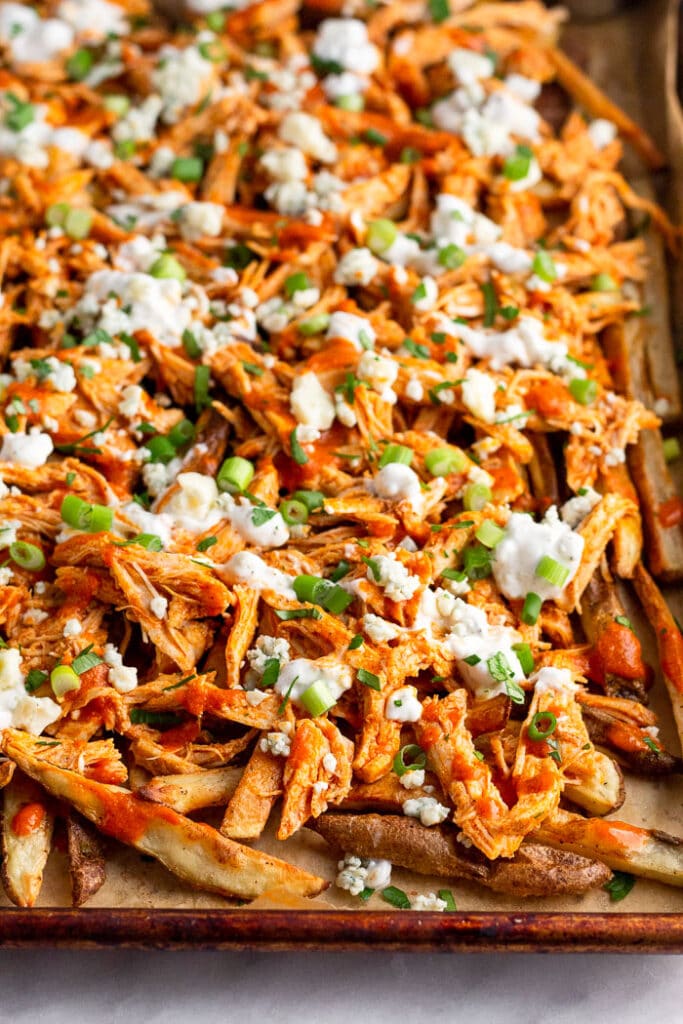 Loaded fries with shredded buffalo chicken, blue cheese, and blue cheese dressing on a baking sheet. They are topped with fresh herbs and green onions.