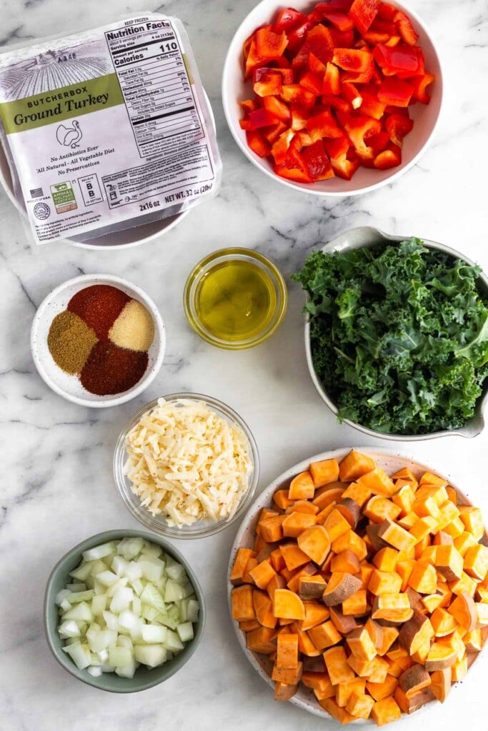 White marble counter with a bowl of diced bell peppers, a bowl of chopped kale, a bowl of diced sweet potatoes, a bowl of diced onions, a bowl of shredded cheese, a small bowl of spices, a bowl of olive oil, and a package of raw ground turkey.