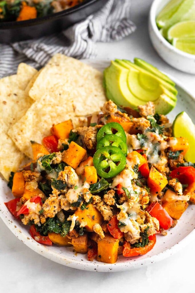 Plate of ground turkey, sweet potato, and veggies topped with melted cheese and sliced jalapeños. Also on the plate are some tortilla chips, sliced avocado, and lime wedges. Behind the plate is a bowl of more lime wedges and a large cast iron.