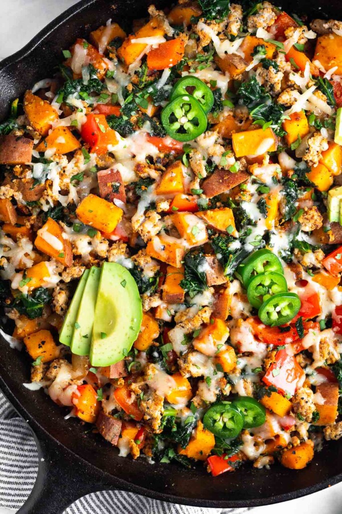 Ground turkey and sweet potato skillet with melted cheese in a large cast iron. It is topped with sliced avocado, jalapeño, and cilantro.