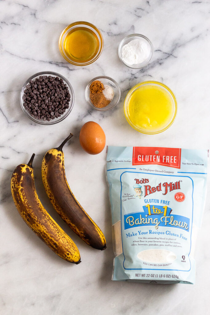 White countertop with a bowl of honey, bowl of baking soda and powder, bowl of melted ghee, package of gluten free flour, two brown bananas, one egg, bowl of spice, and a bowl of chocolate chips.