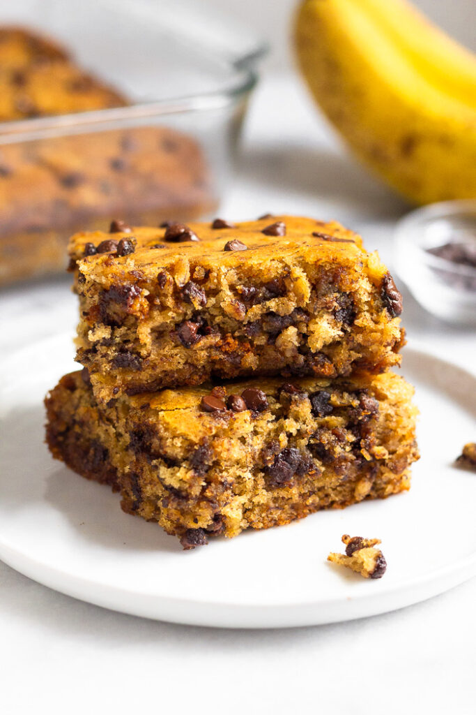 Two healthy banana bread bars on top of each other on a small white plate. Behind them is the rest of the bar in a baking dish and some bananas.