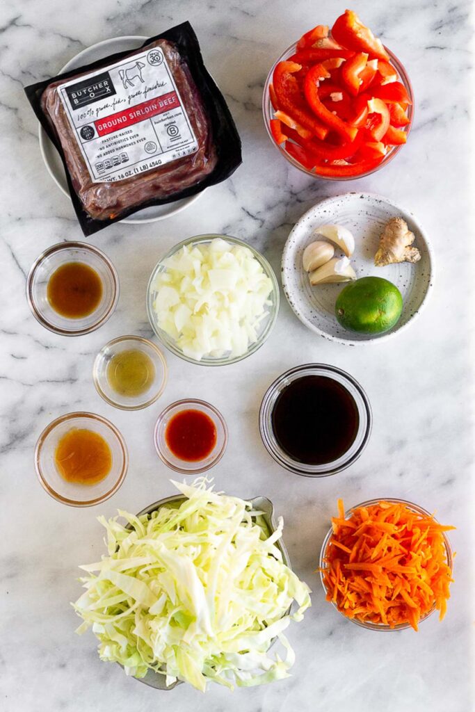 White marble counter with a package of ground beef, a bowl of sliced peppers, a plate of garlic cloves, ginger, and a lime, a bowl of coconut aminos, a bowl of shredded carrots, a bowl of sliced carrots, a bowl of diced onions, and four small bowls of different sauces.