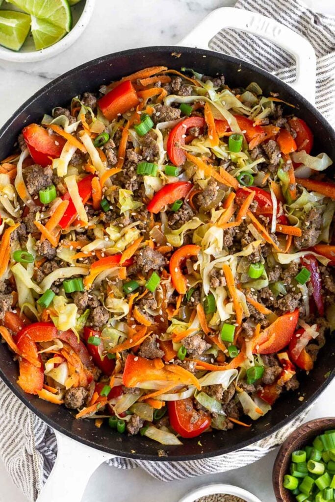 Ground beef and cabbage stir fry in a large cast iron. It is garnished with green onions and sesame seeds. The pan is on top of a kitchen towel with a bowl of lime wedges and a bowl of green onions around it.