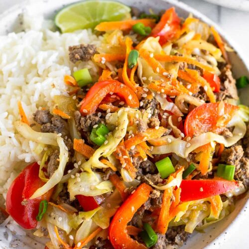 A plate of ground beef and cabbage stir fry with white rice and a lime wedge. It is garnished with sesame seeds and green onions. Behind the plate is another plate and a bowl of sesame seeds.