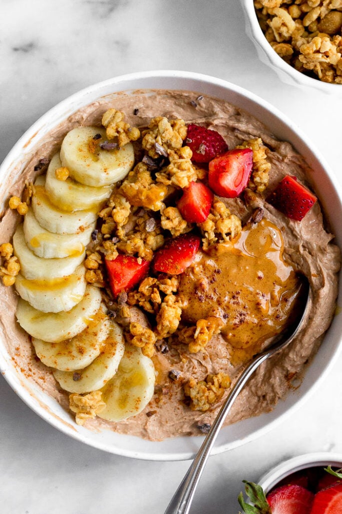 Yogurt bowl topped with sliced banana, peanut butter, granola, strawberries, honey, and cinnamon. A spoon is in the bowl.