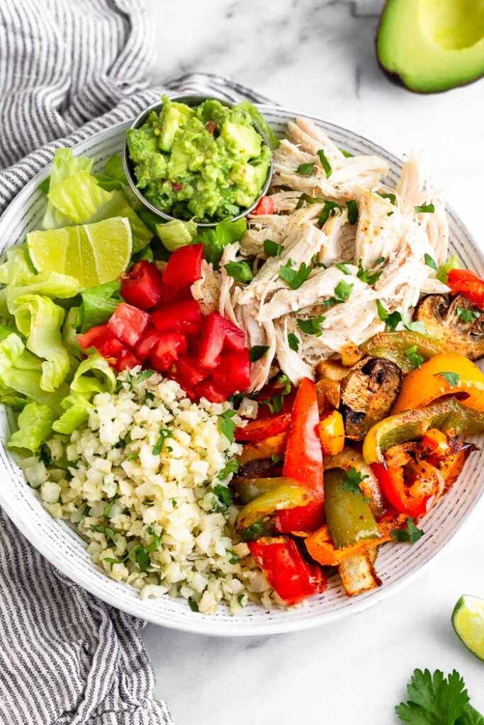 Chicken burrito bowl with shredded chicken, fajita veggies, cauliflower rice, tomato, lettuce, and a small ramekin of guacamole.