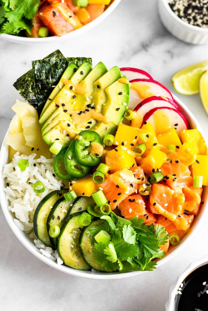 Salmon poke bowl with salmon, cucumbers, rice, avocado, ginger, seaweed, radish, and mango. It is topped with spicy sauce and cilantro.