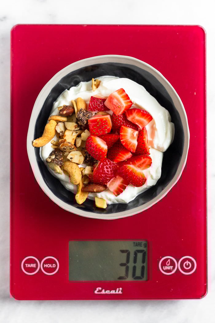 A bowl filled with yogurt, granola, and strawberries on a red food scale to learn how to count macros.