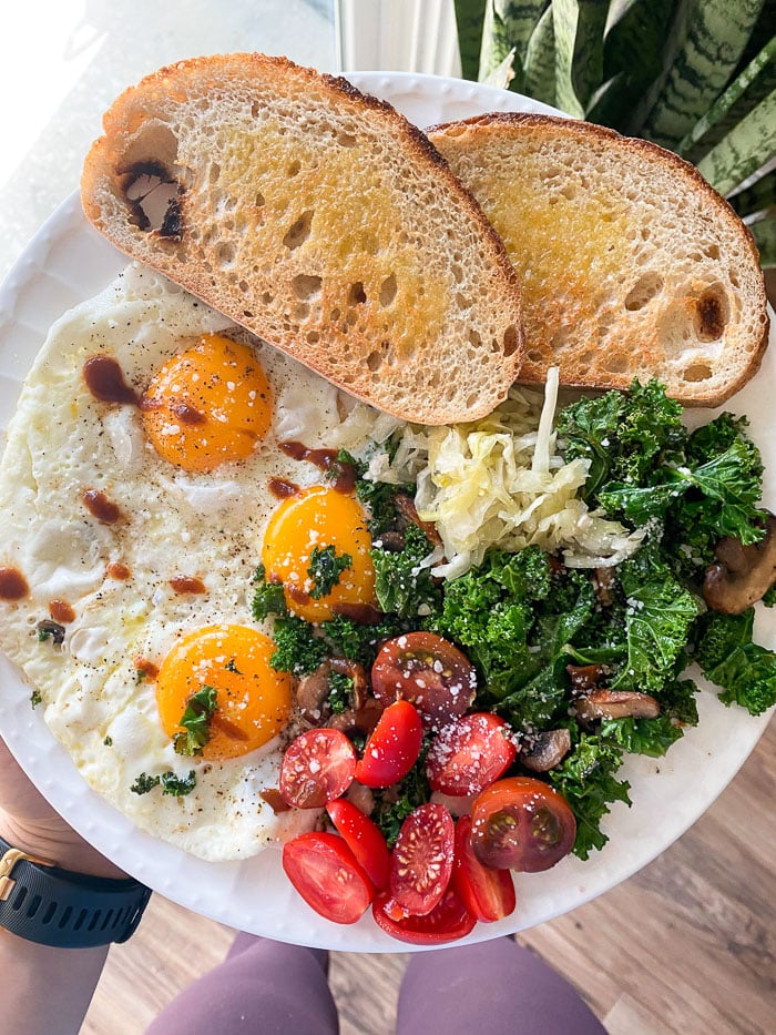 Large plate being held with 3 fried eggs, 2 pieces of toast, veggies, and some sauerkraut. It be being held above a plant.