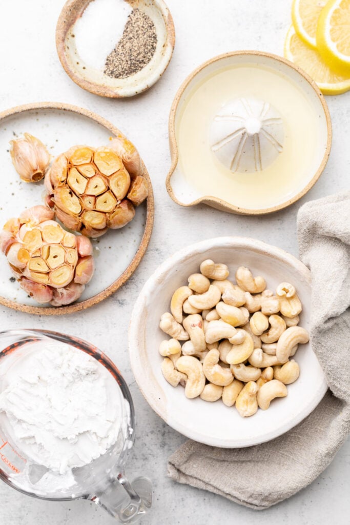 White countertop with a dish of salt and pepper, dish of lemon juice, bowl of cashews, bowl of coconut milk, and a plate of two heads of roasted garlic.