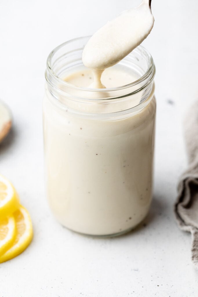 Lemon garlic sauce in a jar with a spoon coming out of it. Next to the jar are some slices of lemon and a tan towel.