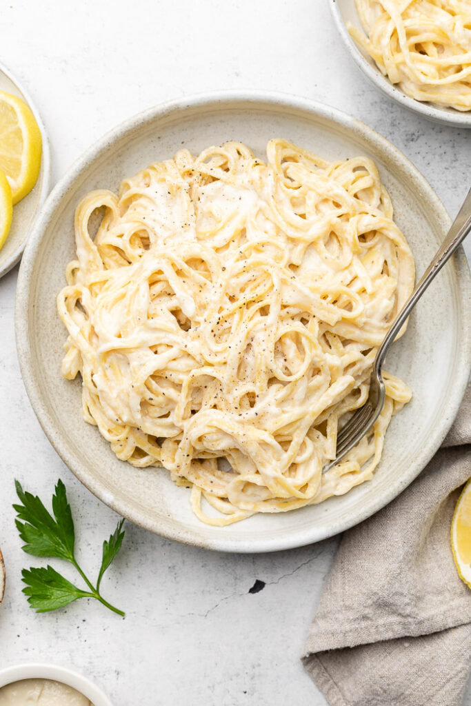 Lemon garlic pasta sauce mixed with pasta in a bowl with a fork in it. Next to it is another bowl, tan towel, parsley, and slices of lemon.