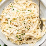 Bowl of pasta with lemon garlic sauce and topped with fresh herbs. A fork is in the pasta and next to the bowl are fresh herbs and slices of lemon.