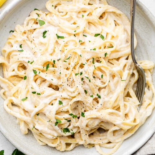 Bowl of pasta with lemon garlic sauce and topped with fresh herbs. A fork is in the pasta and next to the bowl are fresh herbs and slices of lemon.
