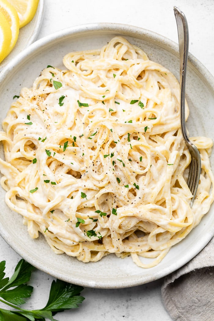 Bowl of pasta with lemon garlic sauce and topped with fresh herbs. A fork is in the pasta and next to the bowl are fresh herbs and slices of lemon.