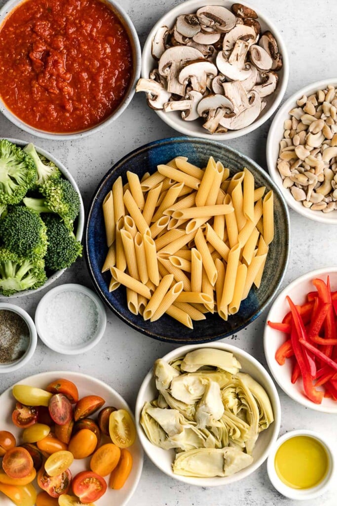 Grey concrete counter with a bowl of sliced mushrooms, a bowl of cashews, a bowl of julienned red bell peppers, a small dish of olive oil, a bowl of quartered artichoke hearts, a bowl of halved cherry tomatoes, a small dish of salt, a small dish of pepper, a bowl of uncooked penne, a bowl of broccoli florets, and a bowl of marinara sauce.