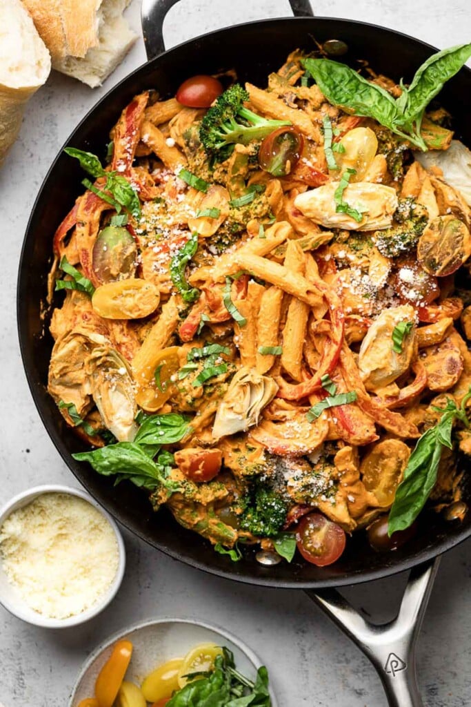 Pasta with veggies tossed with a creamy red sauce in a large skillet. The pasta is garnished with fresh basil and grated cheese. Around the pan is a plate of sliced tomatoes and fresh herbs, a bowl of grated cheese, and some fresh bread.