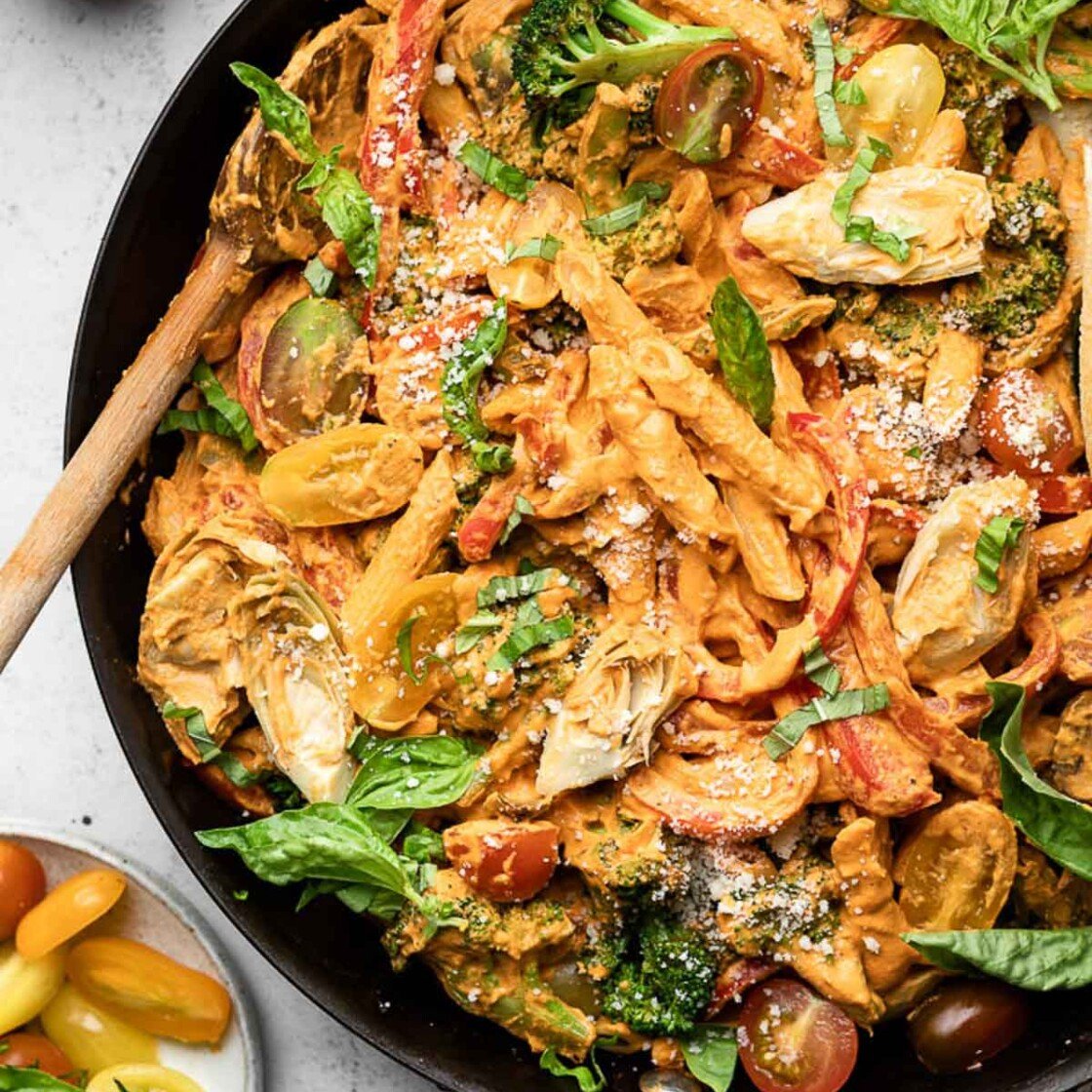 Large skillet filled with vegan pasta with veggies tossed in a creamy red sauce. It is garnished with fresh basil and grated cheese and there is a wooden spoon in the pan. Next to the pan is a plate of fresh basil and halved cherry tomatoes and a bowl of grated cheese.