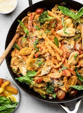 Large skillet filled with vegan pasta with veggies tossed in a creamy red sauce. It is garnished with fresh basil and grated cheese and there is a wooden spoon in the pan. Next to the pan is a plate of fresh basil and halved cherry tomatoes and a bowl of grated cheese.