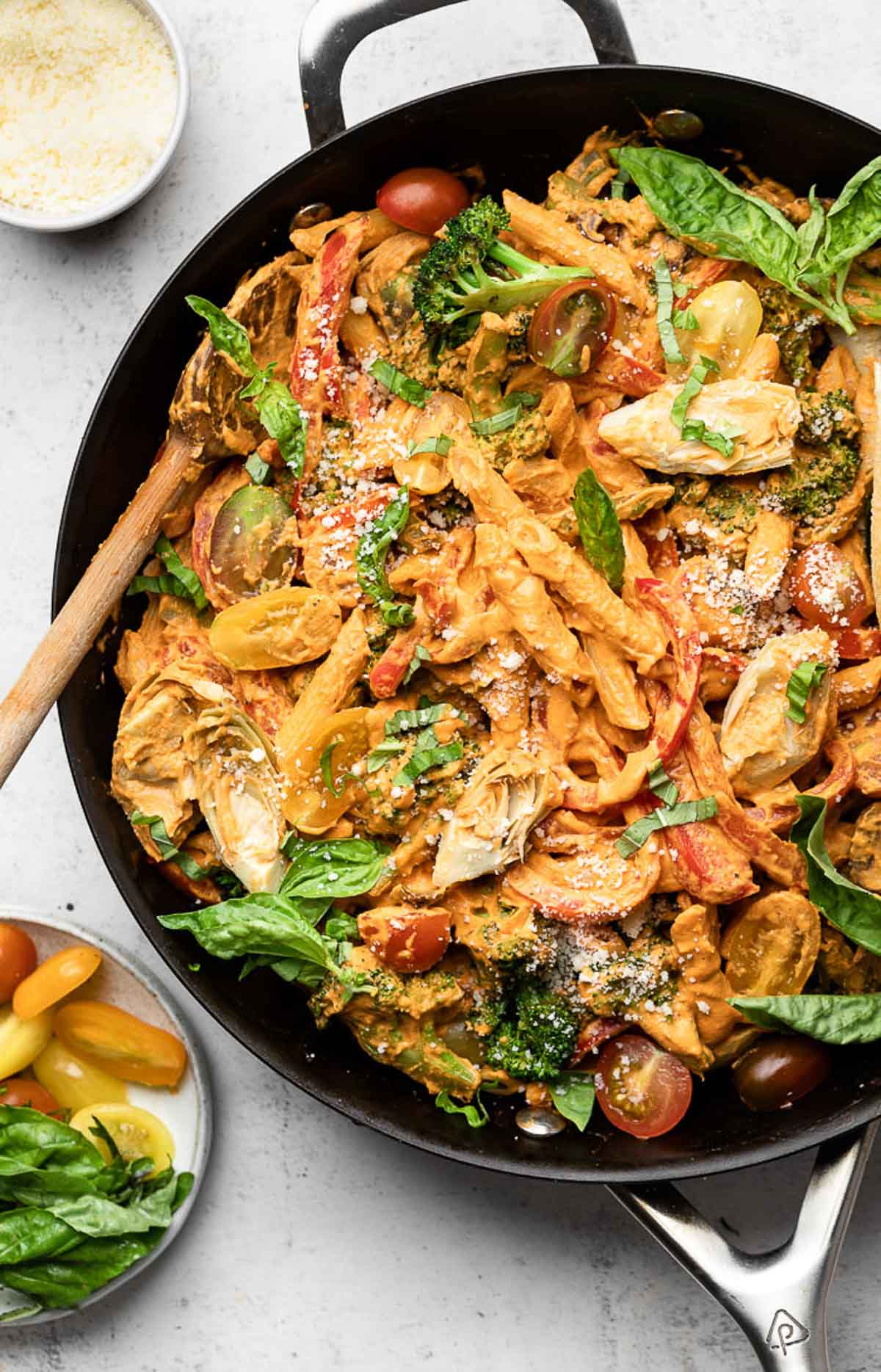 Large skillet filled with vegan pasta with veggies tossed in a creamy red sauce. It is garnished with fresh basil and grated cheese and there is a wooden spoon in the pan. Next to the pan is a plate of fresh basil and halved cherry tomatoes and a bowl of grated cheese.