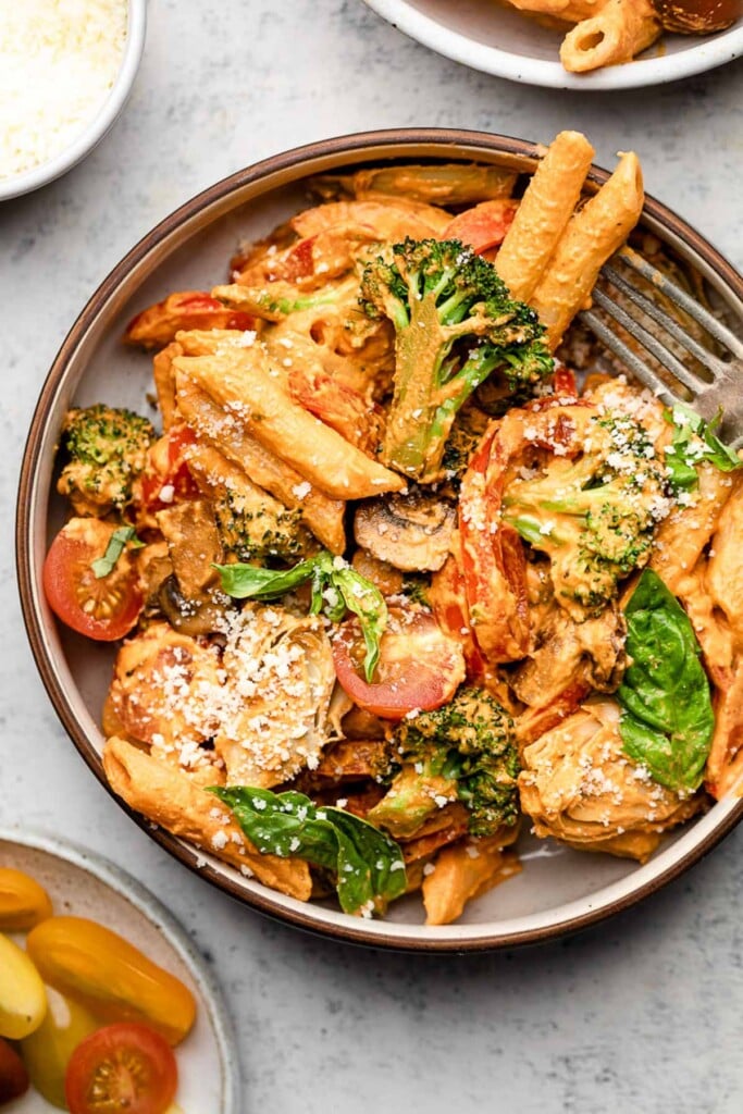 A bowl of creamy vegan pasta with broccoli, peppers, mushrooms, artichoke hearts, and cherry tomatoes. It is garnished with fresh basil and grated cheese. In the bowl is a fork and around the bowl is a plate of halved cherry tomatoes, a bowl of parmesan cheese, and another bowl of pasta.
