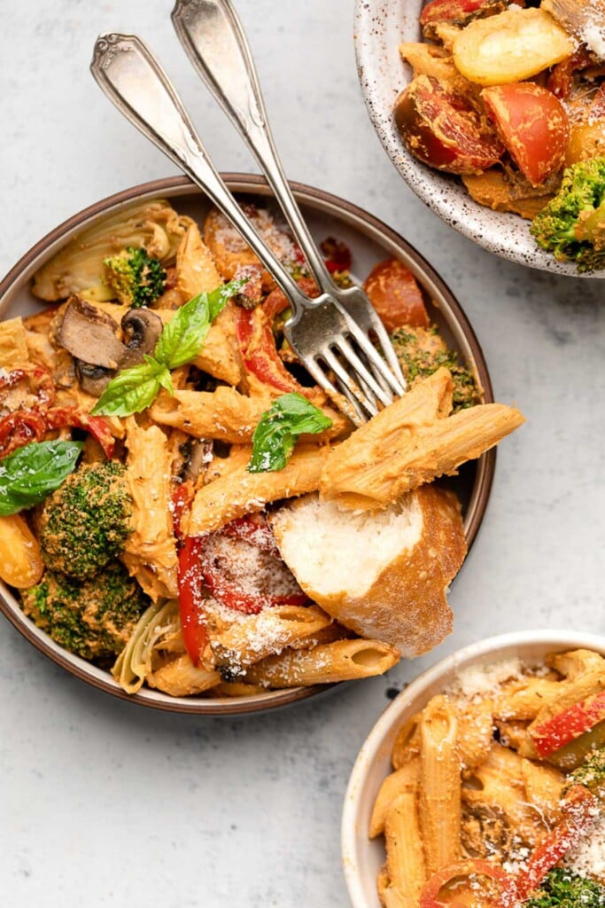 A bowl of pasta with vegetables in a red sauce garnished with basil and parmesan cheese. Also in the bowl is a piece of bread and two forks. Next to the bowl is two more bowls of pasta.